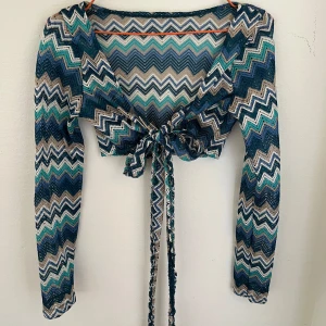 Zigzag mönstrad croppad bolerotopp - Säljer en croppad bolerotopp med långa ärmar och knyt framtill. Toppen har ett färgstarkt zigzag-mönster i blått, turkost, beige och vitt. Materialet är lätt och stretchigt, perfekt för att ge en cool och unik vibe till din outfit.