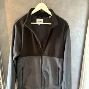Grå fleecejacka från Jack & Jones - Mjuk och skön fleecejacka från Jack & Jones i två nyanser av grått. Jackan har hel dragkedja framtill, hög krage och två fickor med dragkedja. Perfekt för kyliga dagar och enkel att slänga på sig över en hoodie.