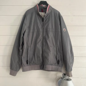 Grå vindjacka från Moncler - Snygg grå vindjacka från Moncler med klassisk bomberkrage och diskret logga på ärmen. Jackan har dragkedja framtill, ribbade muddar och två fickor med dragkedja. Perfekt för dig som gillar stilren och sportig look.