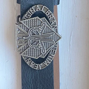 Svart skärp med stor silverspänne - Coolt svart bälte i skinn med ett stort, detaljerat silverspänne där det står 'Denim Jeans Original Brand'. Spännet har en unik form och mönstrad yta som verkligen sticker ut. Perfekt accessoar för att lyfta din outfit med en edgy vibe.