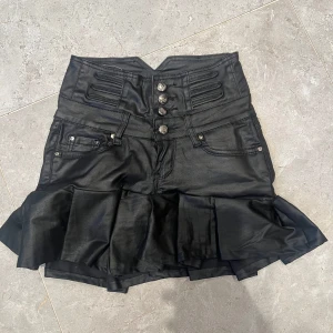 Svart volangkjol i skinnlook - Cool svart kortkjol i skinnimitation med hög midja, dekorativa knappar framtill och volang nertill. Kjolen har fickor både fram och bak samt markerade sömmar för en edgy vibe. Perfekt för dig som vill sticka ut med en unik och trendig stil.