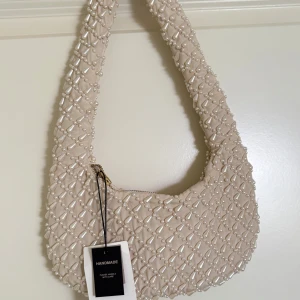 Beige axelväska med pärldetaljer - Superfin beige axelväska från AP Pearls, täckt med glansiga pärlor i ett rutmönster. Väskan har en mjuk, rundad form och stängs med dragkedja. Perfekt för att lyfta vilken outfit som helst med en lyxig touch.