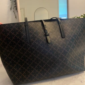 Mönstrad shoppingväska By Malene Birger - Snygg och rymlig shoppingväska från By Malene Birger i svart och brunt med ikoniskt grafiskt mönster. Väskan har långa svarta handtag och justerbar rem, tillverkad i tåligt skinnliknande material. Perfekt för dig som vill ha plats för allt och ändå se chic ut.ena axelremmen behöver lagas då den är limmad. Men den håller nog ett tag till.