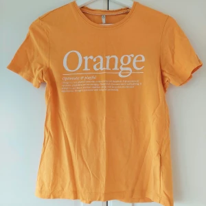Orange t-shirt från ONLY med tryck - Snygg orange t-shirt från ONLY med vit text på bröstet där det står 'Orange'. Klassisk rund halsringning och korta ärmar. Tillverkad i mjuk bomull, perfekt för dig som gillar färg och vill sticka ut lite extra. Så snygg till vita linnebyxor!