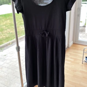 Svart plisserad kortklänning med rosett från Kappahl - Supersöt svart kortklänning med plisserad kjol och liten rosett i midjan. Klänningen har rund halsringning och korta ärmar, vilket ger en klassisk och stilren look. 