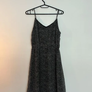 Svart mönstrad sommarklänning - Svart kort klänning med vita prickar och smala axelband. V-ringad framtill och lätt utställd kjol. Väldigt enkel klänning som man kan styla till de mesta. Pris går att diskutera.