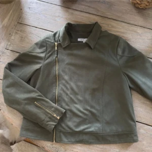 Olivgrön mockajacka från Flash Woman - Snygg olivgrön mockajacka från Flash Woman i bikerstil med sneddragkedja och dragkedjedetaljer vid ärmsluten. Jackan har en klassisk krage och är croppad i modellen, perfekt för att lyfta din outfit med en cool touch.