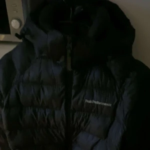 Svart dunjacka Peak Performance - Säljer en svart dunjacka från Peak Performance med huva och dragkedja. Jackan är quiltad och har loggan broderad på bröstet. Perfekt för kalla dagar när du vill hålla värmen och ändå se stilren ut. Barnstorlek. Passar perfekt till någon som är 163cm lång.