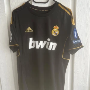 Real Madrid Ronaldo 7 svart bortatröja - Snygg svart Real Madrid bortatröja från Adidas med guldiga detaljer och tryck. Tröjan har korta ärmar, klubbmärke, Champions League-patchar och Ronaldo 7 i guld på ryggen. Materialet är lätt och andas, perfekt för fotboll eller att bära som supporter.