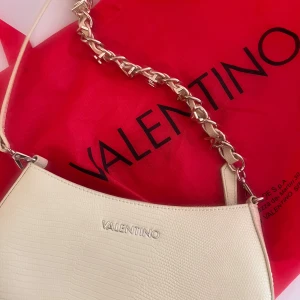 Valentino - Snygg axelväska från Valentino i begie färg med stilren struktur som påminner om lizardmönster. Väskan har en kedjedetalj i guld på axelremmen och dragkedja upptill. Perfekt storlek för det viktigaste och med innerfack för extra organisering.