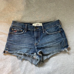Mörkblå jeansshorts från Hollister - Supersnygga mörkblå jeansshorts från Hollister med råa, fransiga kanter och klassisk femficksdesign. Shortsen har låg midja och slitna detaljer. 
