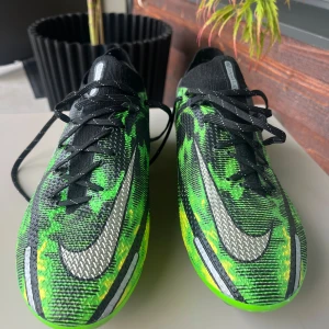Nike Phantom GT2 Elite fotbollsskor - Säljer ett par Nike Phantom GT2 Elite fotbollsskor i svart, grönt och gult med silverfärgade detaljer. Skorna har snörning, mönstrad ovandel i syntet och en platt sula med dobbar för bästa grepp på planen. Perfekta för dig som vill sticka ut på matchen.