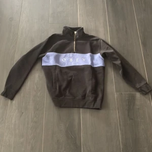 Svart half zip tröja från Obey - Svart tröja från Obey med en ljusblå panel över bröstet och vit Obey-logga. Half zip-dragkedja och hög krage ger en chill vibe. Långärmad och tillverkad i mjuk bomull, perfekt för lager-på-lager. Snygg streetwear som funkar året runt.