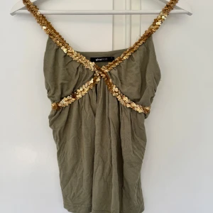 Olivgrön linne med guldpaljetter Gina Tricot - Supersnygg olivgrön linne från Gina Tricot med breda axelband täckta av glittrande guldpaljetter. Toppen har en tight passform över bysten me lösare drapering runt midjan. 