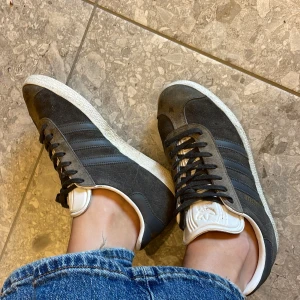 Adidas Gazelle grå mockasneakers - Säljer ett par klassiska Adidas Gazelle sneakers i mörkgrå mocka med svarta ränder och vit sula. Skorna har snörning, rund tå och vit plös med Adidas-logga