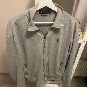 Grå ziptröja Polo Ralph Lauren M - Snygg grå ziptröja från Polo Ralph Lauren i bomull med klassisk krage och dragkedja framtill. Tröjan har två fickor med dragkedja och den ikoniska blå loggan broderad på bröstet. Pris kan diskuteras