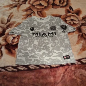 Miami BAPE x Inter Miami fotbollströja - Snygg limited edition fotbollströja från BAPE x Inter Miami med grått och vitt camouflage-mönster. Stor MIAMI-print på bröstet, BAPE-logga och klubbmärken på fram- och ärm. Baksidan har BAPE och siffran 93 i svart. Tillverkad i mjuk bomull.