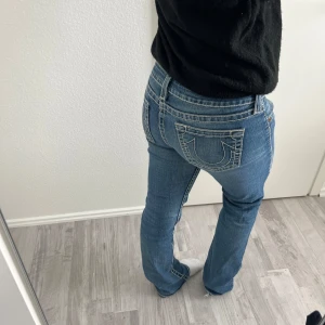 True Religion blå bootcut jeans - Säljer ett par blå jeans från True Religion med tydliga vita kontrastsömmar och ikoniska bakfickor. Modellen har låg midja och bootcut-ben. Har hål vid innerlåren men syns inte när man har på sig dom. Innerbebslängd 80