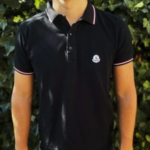 Svart Moncler pikétröja med röd/vit kant - Svart piké från Moncler med klassisk krage och korta ärmar. Tröjan har Moncler-logga på bröstet och snygga rödvita detaljer på krage och ärmslut. Tillverkad i mjuk bomull som är skön mot huden. Perfekt för dig som gillar stilren och sportig look.                                                                                                                                         Nypris: 3600kr GORILLAPRIZE:1520kr            Skick:8/10 mycket fint skick         KVITTO MEDKOMMER     