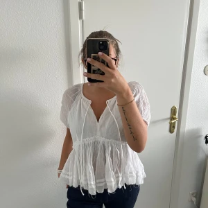 Vit blus med volang från Gina Tricot - Supersöt vit blus från Gina Tricot med korta puffärmar, v-ringning och knytband fram. Blusen har volangkant och är tillverkad i tunn, luftig bomull som ger en chill och flowy vibe. 