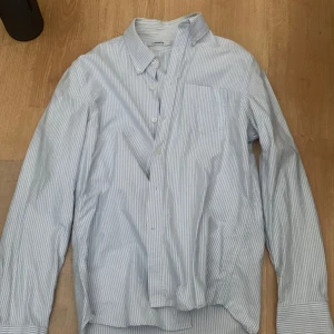 Randig skjorta från J.Lindeberg - Snygg skjorta från J.Lindeberg i vitt och ljusblått med tunna ränder. Klassisk krage, bröstficka och knäppning framtill. Perfekt för dig som gillar stilrena och fräscha plagg. Skjortan är långärmad och i mjuk bomull.