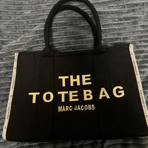 Svart tote bag från Marc Jacobs - Säljer en svart tote bag från Marc Jacobs med tydlig logga framtill och cool textdetalj på baksidan, har både handtag och axelrem med Marc Jacobs-tryck samt dragkedja upptill. Perfekt för dig som vill ha en rymlig och snygg väska.