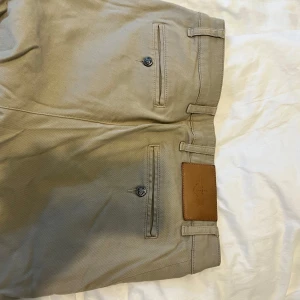 Beige chinos med raka ben - Snygga beige chinos med raka ben och klassiska fickor både fram och bak. Byxorna har bälteshällor och en stilren läderpatch bak i midjan. Perfekta för en clean och avslappnad look. Tillverkade i mjuk bomull för skön känsla hela dagen.