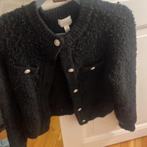 Svart kofta från H&M med silvriga knappar - Säljer en svart, fluffig kofta från H&M i storlek S. Koftan har silvriga dekorativa knappar framtill och två fickor på bröstet. Den är stickad och har lång ärm samt en klassisk rund halsringning. Perfekt för lager-på-lager och snygg till jeans.