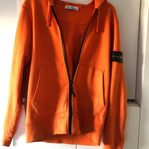 Stone Island Garment Dyed Zip Hooded Sweatshirt  - Snygg orange hoodie från Stone Island med dragkedja och klassisk logga på ärmen. Tröjan har huva med snörning, två stora fickor fram och är tillverkad i mjuk bomull. Perfekt för dig som vill sticka ut med färg och streetwear-vibe.