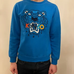 Blå Kenzo sweatshirt med tigertryck - Säljer en blå sweatshirt från Kenzo med ikoniskt broderat tigerhuvud och färgglada bokstäver på bröstet. Tröjan har rund hals, långa ärmar och ribbade muddar. Perfekt för dig som vill sticka ut med en streetstyle-look.