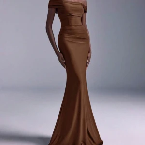 Brun offshoulder långklänning - Supersnygg brun långklänning med offshoulder-design och figurnära passform. Klänningen har en slät yta och markerad midja, vilket ger en elegant siluett. Materialet har en lätt glans och tyget faller mjukt ner mot golvet. Perfekt för dig som vill sticka ut med stil.