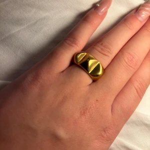 Guldig chunky ring med pyramidform - Säljer en chunky ring i guld med två coola pyramidformade detaljer på ovansidan. Ringen har en bred och blank yta som verkligen sticker ut och ger en edgy vibe. Perfekt för dig som gillar statement-smycken och vill ha något som syns.