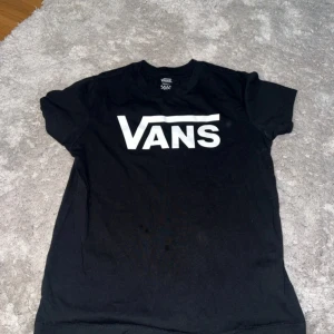 Svart Vans t-shirt med logga - Svart t-shirt från Vans i storlek small med stor vit Vans-logga på bröstet. Klassisk rund hals och korta ärmar. Materialet är mjuk bomull som känns skönt mot huden. Perfekt för dig som gillar streetwear och enkel stil.