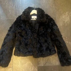 Svart pälsjacka från Vila, stl 34 - Mysig svart pälsjacka från Vila med fluffig känsla och klassisk krage. Jackan har långa ärmar och stängs med knappar framtill. Perfekt för dig som vill ha en statementjacka med lyxig look och skön värme under kyliga dagar.