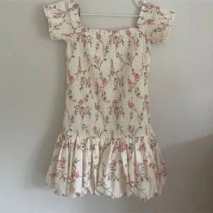 Blommig kortklänning med volang - Supersöt kortklänning med blommigt mönster i rosa, gult och grönt på vit botten. Klänningen har puffiga korta ärmar, smockad överdel och en bred volang nedtill som ger extra flärd. Perfekt för sommarens alla tillfällen! Pris kan diskuteras 