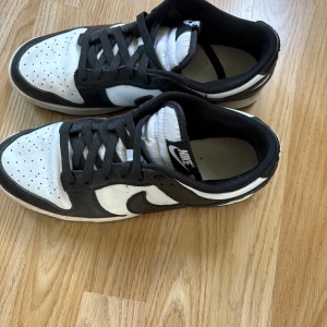 Nike Dunk Low svart/vit sneakers - Snygga Nike Dunk Low sneakers i svart och vitt med klassisk design och lågt skaft. Skorna har svart snörning, vit tåbox med perforeringar och svart yttersula. Perfekta för dig som gillar streetstyle. Skorna är i ganska bra skick.