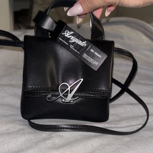 Svart minihandväska Axel Arigato - Säljer en svart minihandväska från Axel Arigato med stilren design och silverfärgad A-logga framtill. Väskan har ett smidigt axelband och är gjord i slätt skinn. P