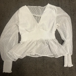 Vit blus med v-ringning och volang - Superfin vit blus med v-ringning, transparenta långärmar och smock i midjan och vid ärmsluten. Blusen har små prickar i tyget och volang nertill. Säljer den då den är lite för stor på mig. Köpt second hand så vet tyvärr inte vart den är ifrån💞
