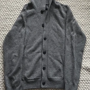 Grå cardigan med luva - Mysig grå kofta med huva och stora svarta knappar framtill. Tröjan har långa ärmar, ribbade muddar och två fickor på sidorna. Perfekt för lager-på-lager och chill dagar. Enkel och stilren design som funkar till det mesta.