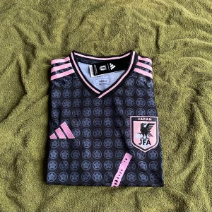 Japan Adidas fotbollströja svart/rosa L - Japan fotbollströja från Adidas i svart med rosa detaljer och mönster. Tröjan har korta ärmar, V-ringning och ett unikt blommönster med katana-tryck på framsidan. Klassiska Adidas-ränder på axlarna och JFA-emblem på bröstet. Tillverkad i lätt och ventilerande material.