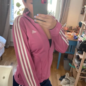 Rosa Adidas ziptröja med ränder - Säljer en rosa Adidas ziptröja med klassiska vita ränder längs ärmarna och vit logga på bröstet. Tröjan har hög krage, dragkedja hela vägen och långa ärmar. Perfekt för en sportig look eller chill vardagsstil.