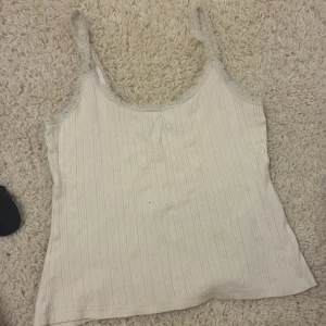 Vit spetskantad linnetopp 157 XS - Supersöt vit linnetopp från 157 i storlek XS. Toppen har tunna axelband och är dekorerad med spets längs kanterna. Tyget har ett diskret randigt mönster och små broderade detaljer som ger en romantisk vibe. Perfekt att styla med jeans eller kjol.