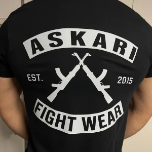 Svart Askari Fight Wear t-shirt - Svart t-shirt från Askari Fight Wear med stor vit logga och korsade gevär på ryggen samt mindre tryck på bröstet. Klassisk rund hals och korta ärmar. Skön passform och tillverkad i mjuk bomull, perfekt för dig som gillar streetwear och träningsstil.