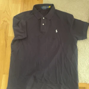 Polo Ralph Lauren pikétröja mörkblå - Klassisk svart pikétröja från Polo Ralph Lauren med vit broderad logga på bröstet. Tröjan har korta ärmar, krage och knäppning med två knappar. Tillverkad i mjuk bomull som är skön mot huden. Perfekt för en clean och stilren look.