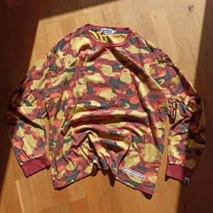 A Bathing Ape camo långärmad tröja - Äkta. Säljer en långärmad tröja från A Bathing Ape i storlek 2XL med ikoniskt camo-mönster i gul, röd, orange och svart. Tröjan har ribbade muddar och rund halsringning i vinröd färg. Stor BAPE-logga på ryggen och klassisk BAPE-tag på ärmen. Perfekt för streetwear-stilen. Mer information, bilder och prisdiskutering i dm. Passar M,L, XL.