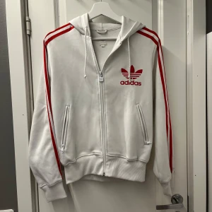 Vit Adidas hoodie med röda ränder - Säljer en vit Adidas hoodie med röda ränder längs ärmarna och stort rött Adidas-tryck på bröstet. Tröjan har huva med snörning, dragkedja framtill och två fickor med dragkedja. Klassisk streetstil och skön passform, perfekt för retrostil eller vilken stil som helst.
