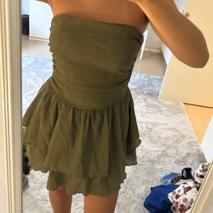 Grön axelbandslös kortklänning - Säljer en snygg grön axelbandslös kortklänning med volang i kjolen. Klänningen har en smockad överdel som sitter tight och ger en cool siluett. Perfekt för dig som gillar en enkel men trendig look. Materialet känns lätt och luftigt, perfekt för varma dagar.