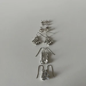 Set med fem par silvriga örhängen - Säljer ett set med fem par silvriga örhängen i olika modeller. Inkluderar små studs med stenar, cirkelformade örhängen med glittriga detaljer, droppformade hängen och större örhängen med bladformade stenar. 