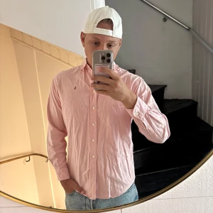 Rosa skjorta från Ralph Lauren - Säljer en klassisk rosa skjorta från Ralph Lauren med button-down krage och vita knappar. Skjortan har lång ärm och den ikoniska broderade loggan på bröstet. Perfekt för dig som gillar stilrena och tidlösa plagg. Små slitage som knappt märks 