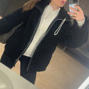 Svart teddyjacka med dragkedja - Mysig svart teddyjacka med oversized passform och dragkedja framtill. Jackan har en cool pärldetalj vid kragen och är perfekt för kalla dagar. Långärmad och fluffig, passar dig som gillar streetstyle och vill hålla dig varm.